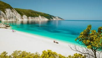 Fteri beach lagoon, Cephalonia Kefalonia, Greece. Tourists under umbrella relax near clear blue emerald turquise sea water. White rocks in background.