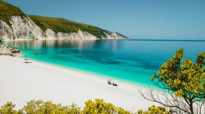 Fteri beach lagoon, Cephalonia Kefalonia, Greece. Tourists under umbrella relax near clear blue emerald turquise sea water. White rocks in background.